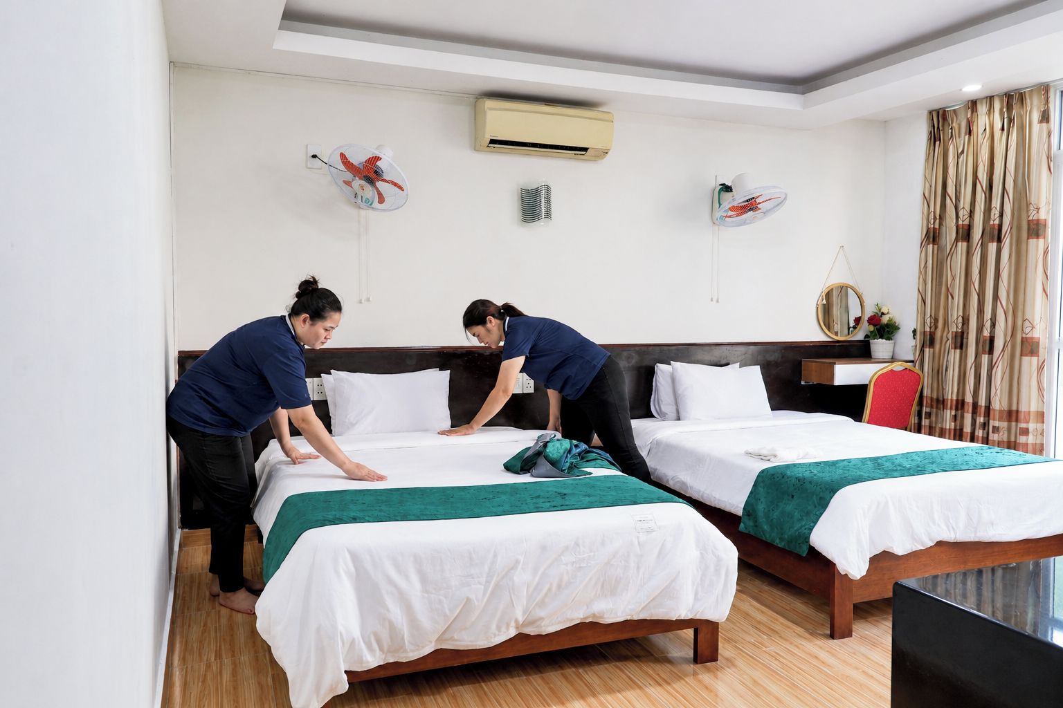 A hotel room with two beds, a desk, and a chair. There are wall-mounted fans and an air conditioning unit.