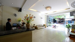 A hotel reception area with a receptionist, a reception desk, a sofa, a coffee table, and a large potted plant. - Catba Island Hotel (Hai Phong)