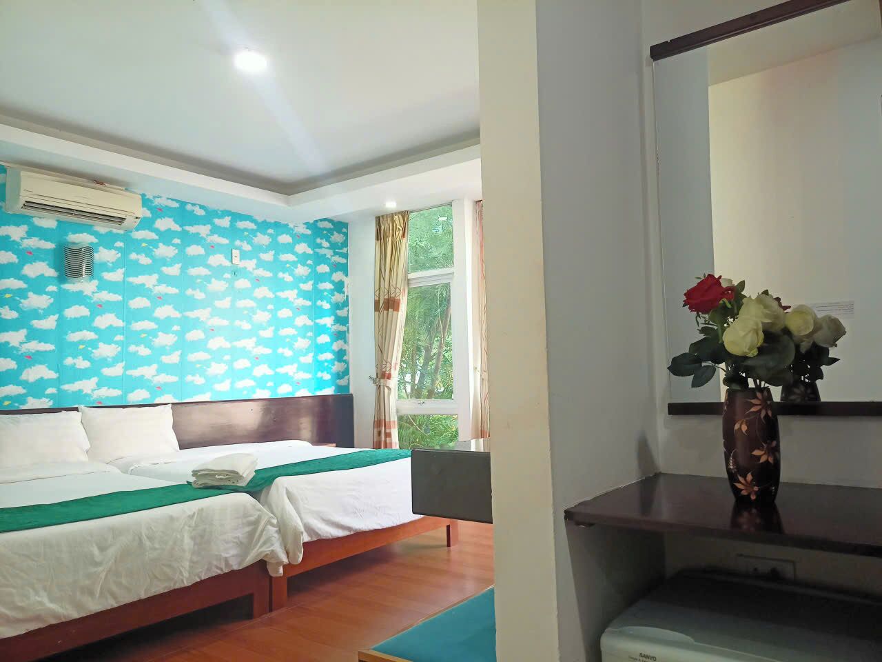 Habitación de hotel con dos camas, un ventanal con cortinas, una pared con diseño de nubes y un jarrón con flores sobre una mesa.