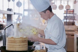 Ein Koch in weißer Uniform bereitet Essen in einer Küche mit Dampfgarer und Wok zu.