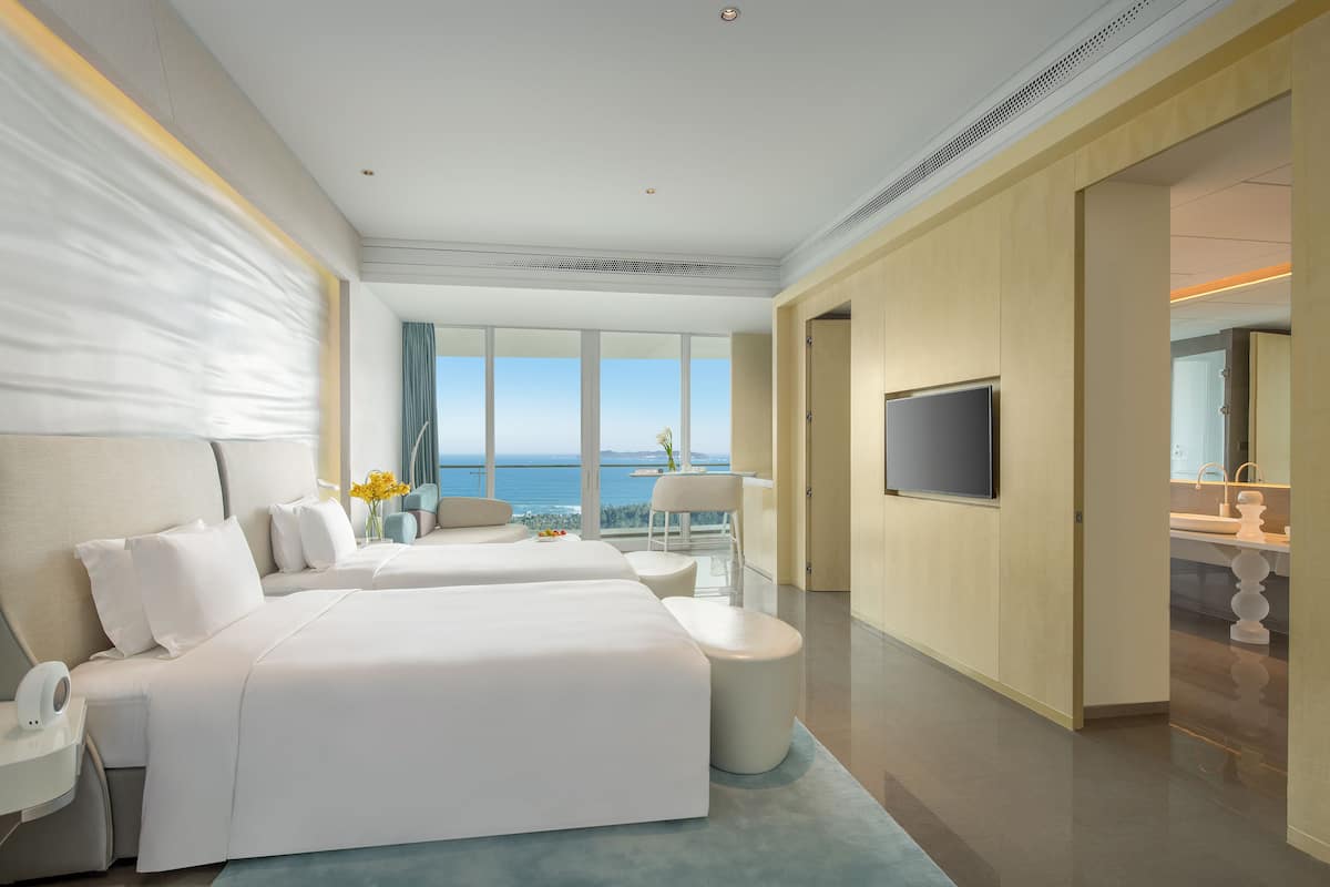 Habitación de hotel con dos camas, televisión, ventana grande con vistas al mar y balcón con una silla y una mesa.