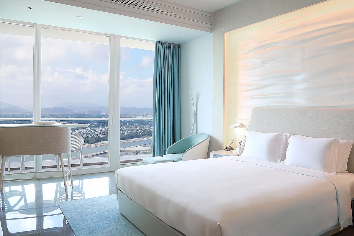 Habitación de hotel con una cama grande, una silla, una mesita de noche y vistas a un paisaje urbano a través de un ventanal amplio.