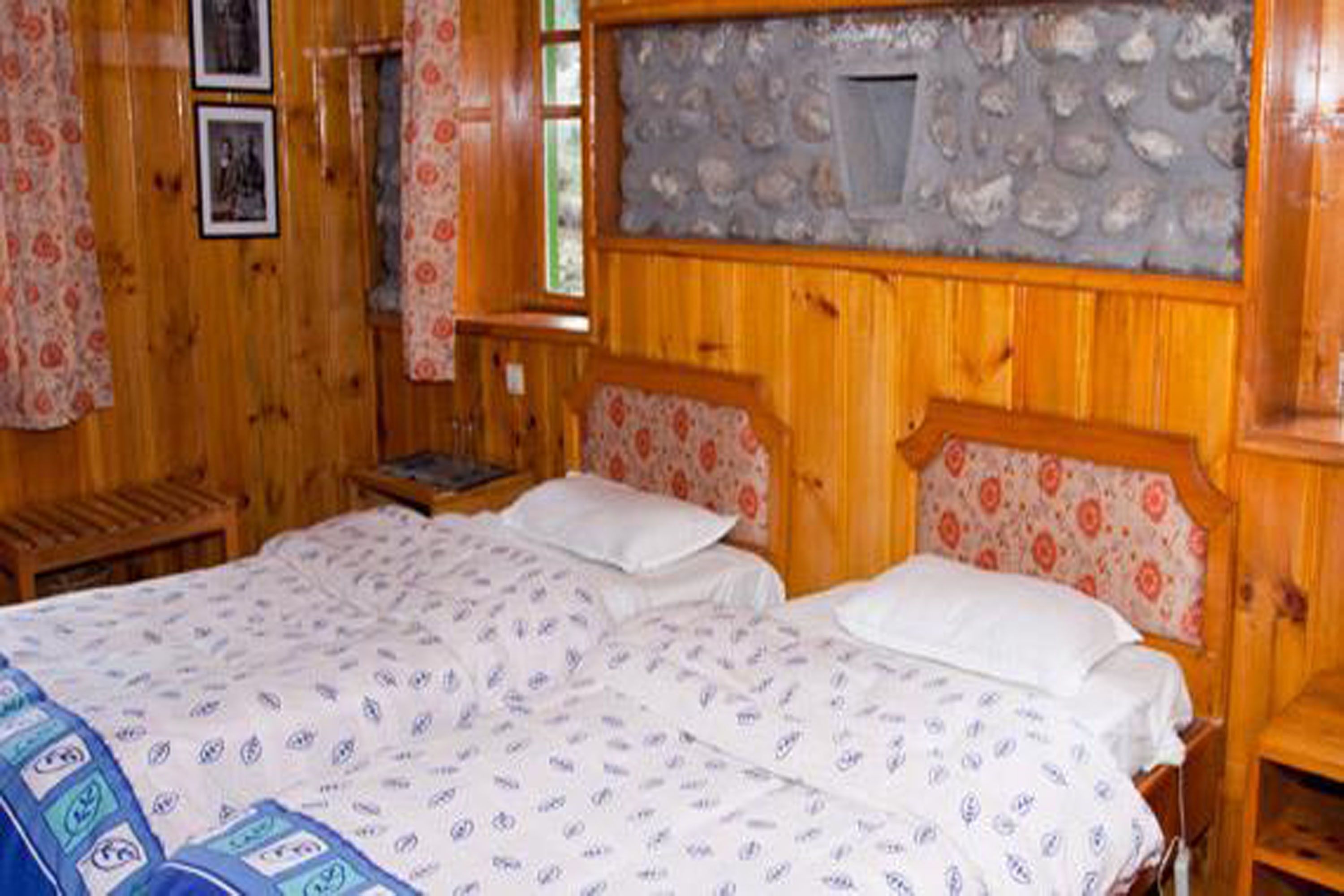 A wooden room with two beds, floral-patterned curtains, and a stone wall with a small window.