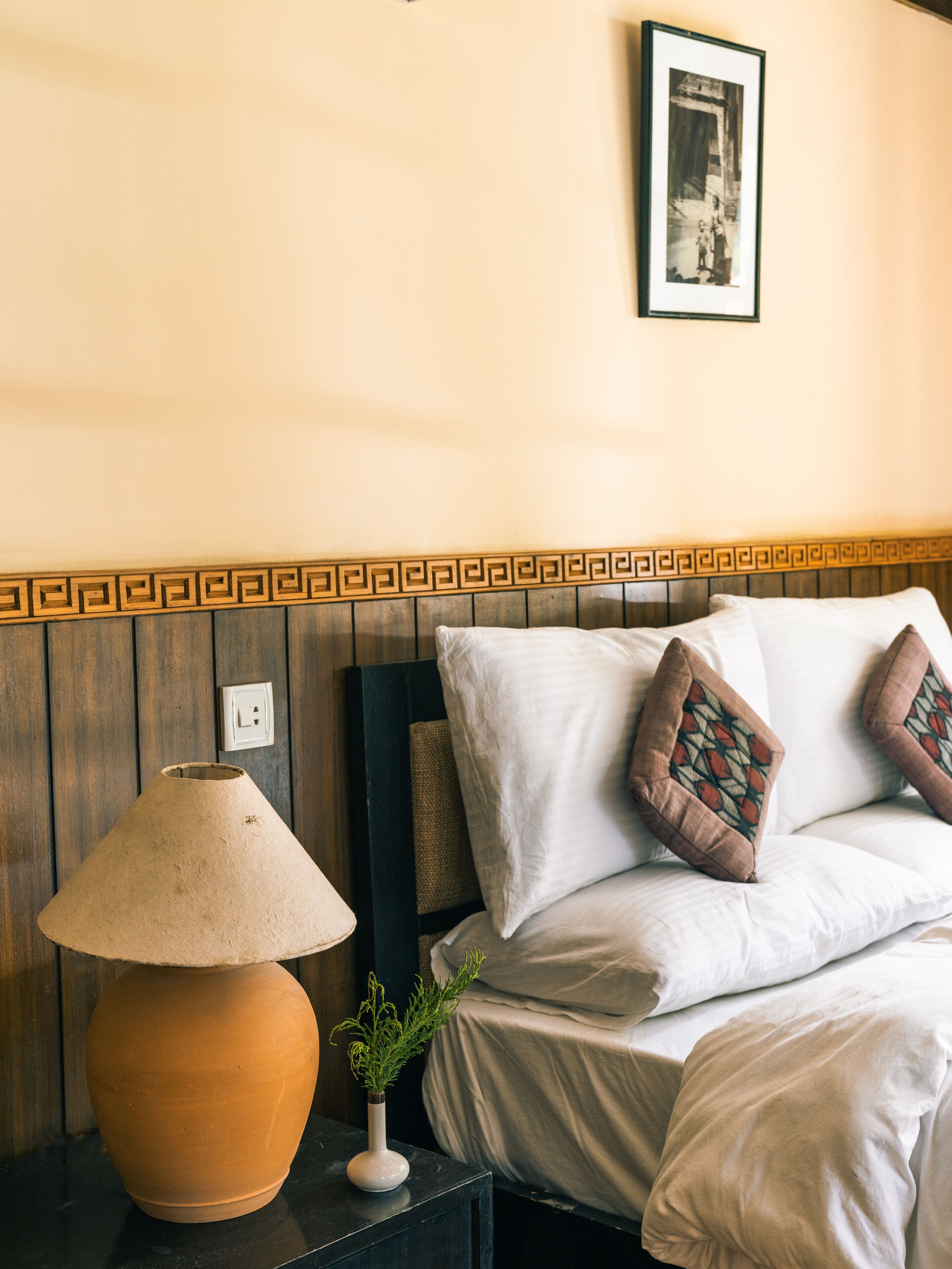 Un lit avec du linge de lit blanc et des coussins à motifs, une lampe beige sur une table de chevet et une photo encadrée accrochée au mur.