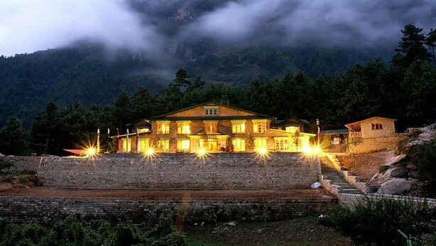 A stone building with multiple windows and a staircase leading up to it, surrounded by trees and mountains.