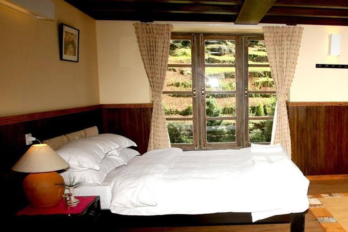 A bedroom with a bed, a nightstand with a lamp, a window with curtains, and a view of greenery outside.