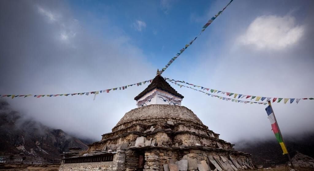 A traditional stupa with a stone base and a pointed roof, surrounded by mountains and clouds.