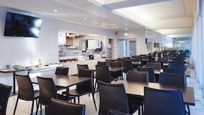 Un espace repas moderne avec plusieurs tables et chaises, une cuisine en arrière-plan et un téléviseur à écran plat fixé au mur.