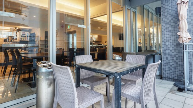Un espace de restauration en plein air avec des chaises en osier et une table en bois, des cloisons en verre et un mur recouvert de carreaux.