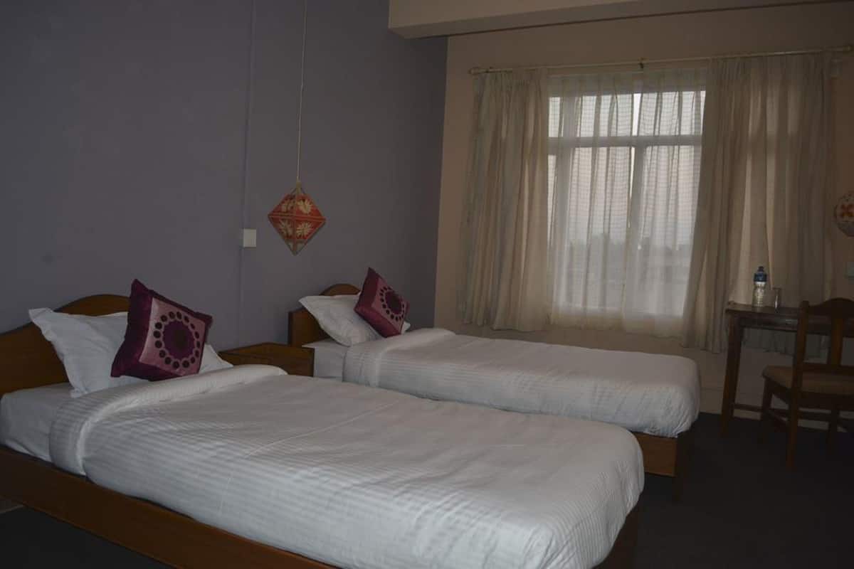 Habitación de hotel con dos camas individuales, cabecero de madera, una mesita con una silla y una ventana con cortinas translúcidas.