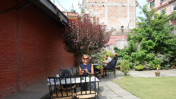 An outdoor patio area with tables and chairs, a brick wall, and a small garden with a tree and a potted plant. - Bodhi Guest House (Kathmandu)