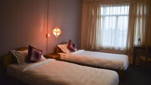 A hotel room with two single beds, a nightstand, a table, and a window with curtains. - Bodhi Guest House (Kathmandu)