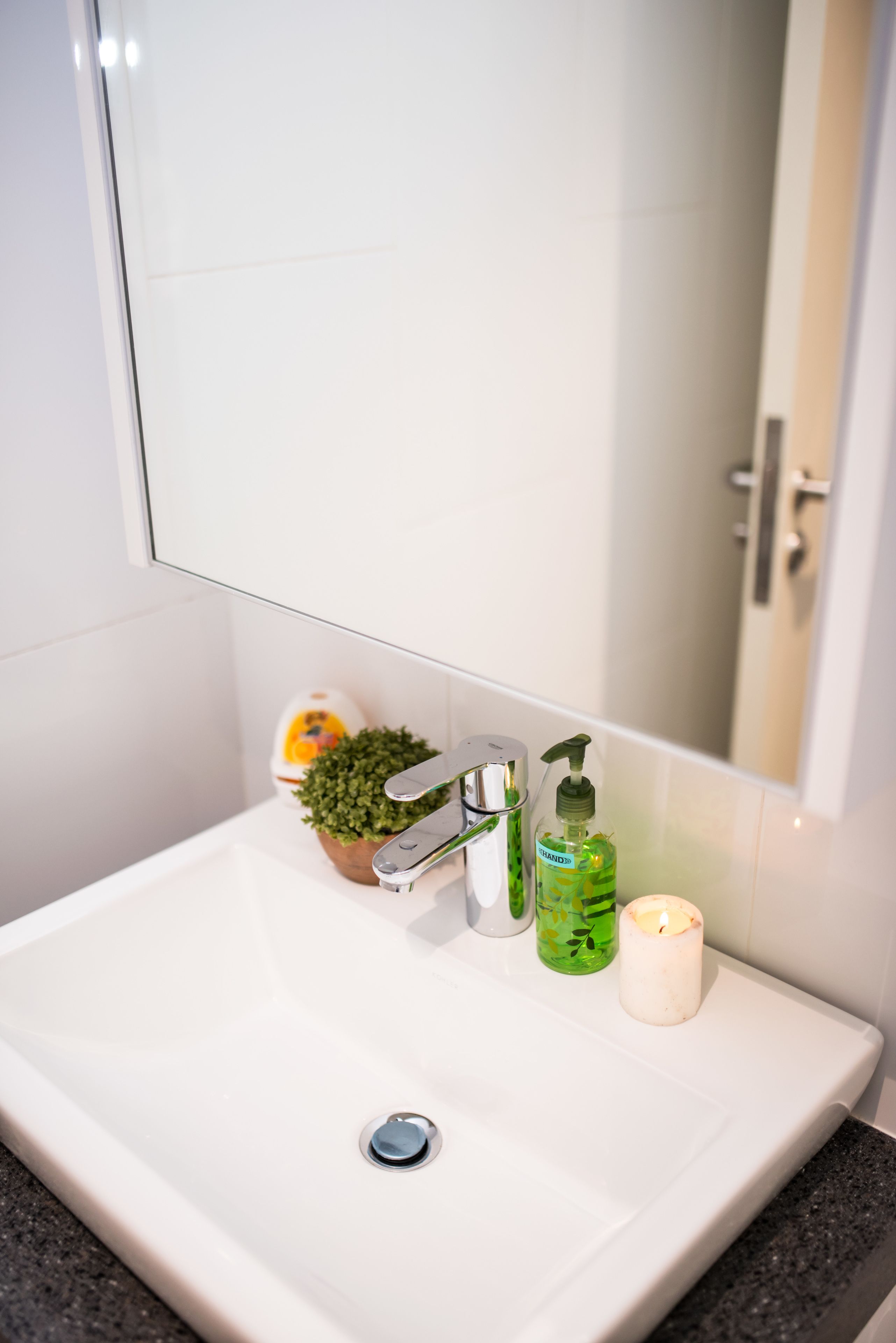 A bathroom sink with a mirror, a green soap dispenser, a small potted plant, and a lit candle.