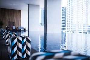 An indoor infinity pool with a view of a tall building, lounge chairs, and a wooden wall.
