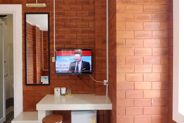 A bathroom with a television mounted on a brick wall, a mirror, a sink with a trash bin, and a toilet. - Changsova Hotel (Ban Chang)