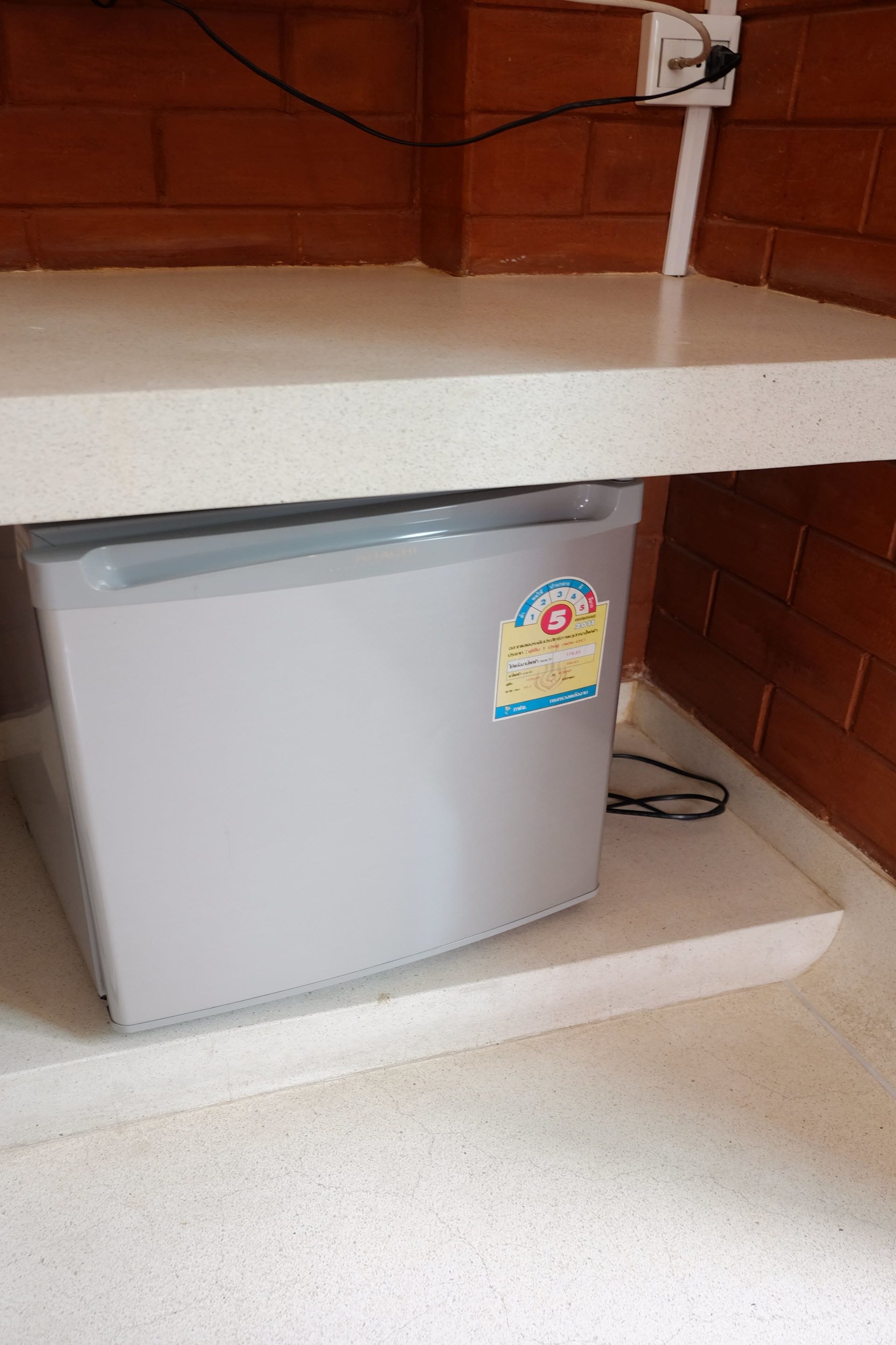 Un petit réfrigérateur placé sous un plan de travail blanc, avec une prise électrique sur le mur.