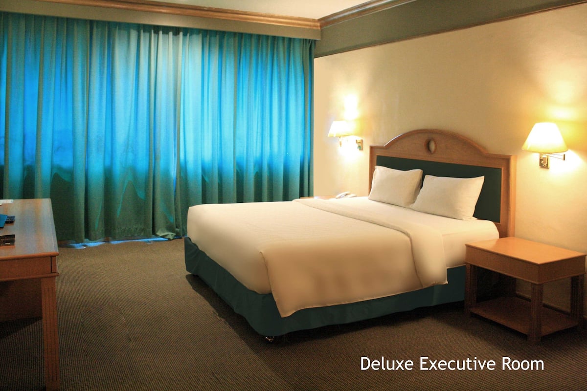 A Deluxe Executive Room with a large bed, two bedside lamps, a wooden nightstand, and a view of the outside through curtains.