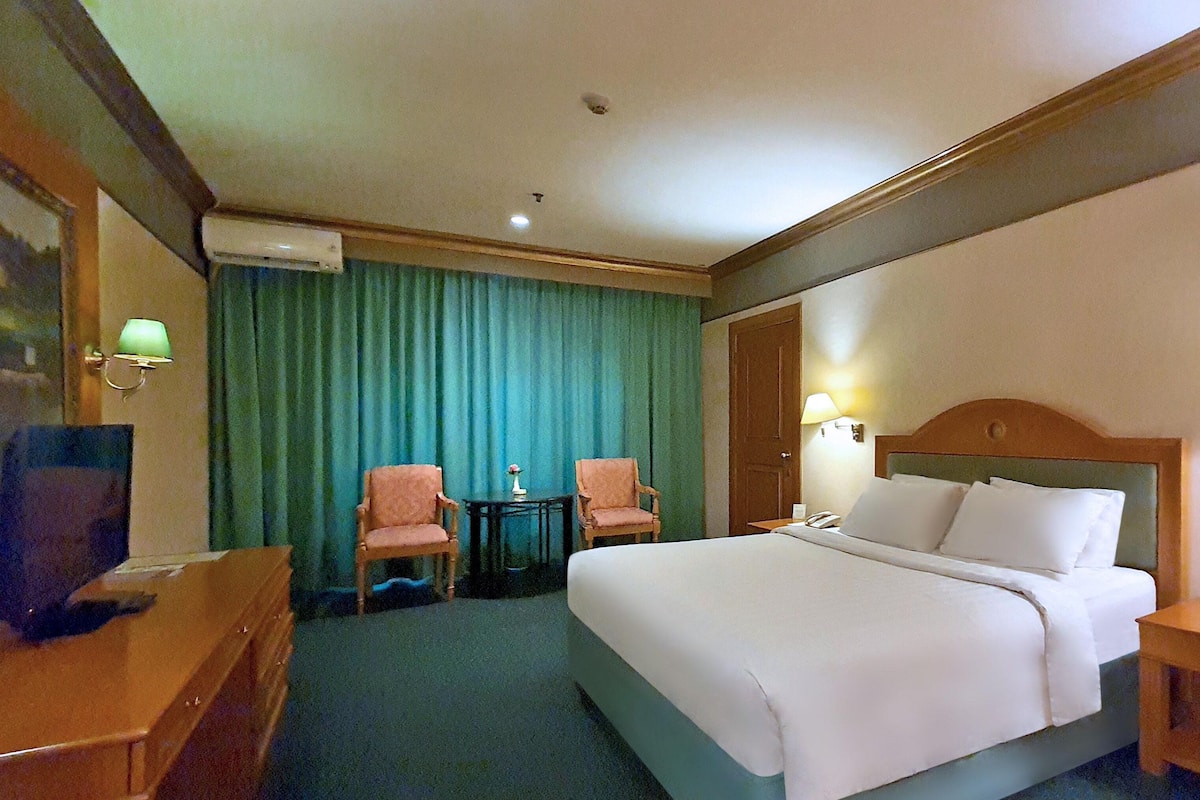 A hotel room with a bed, two chairs, a desk, a lamp, and a television.