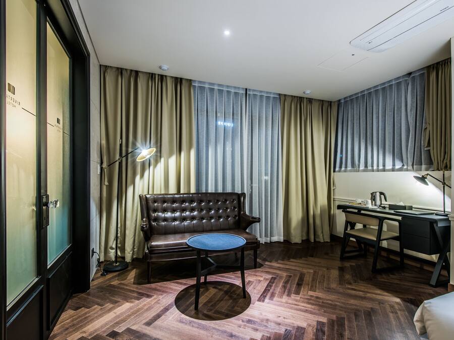 A hotel room with a leather sofa, a round table, a desk with a chair, and a large window with curtains.