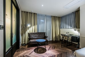 Habitación de hotel con sofá de cuero, una mesa redonda, un escritorio con silla y un amplio ventanal con cortinas.