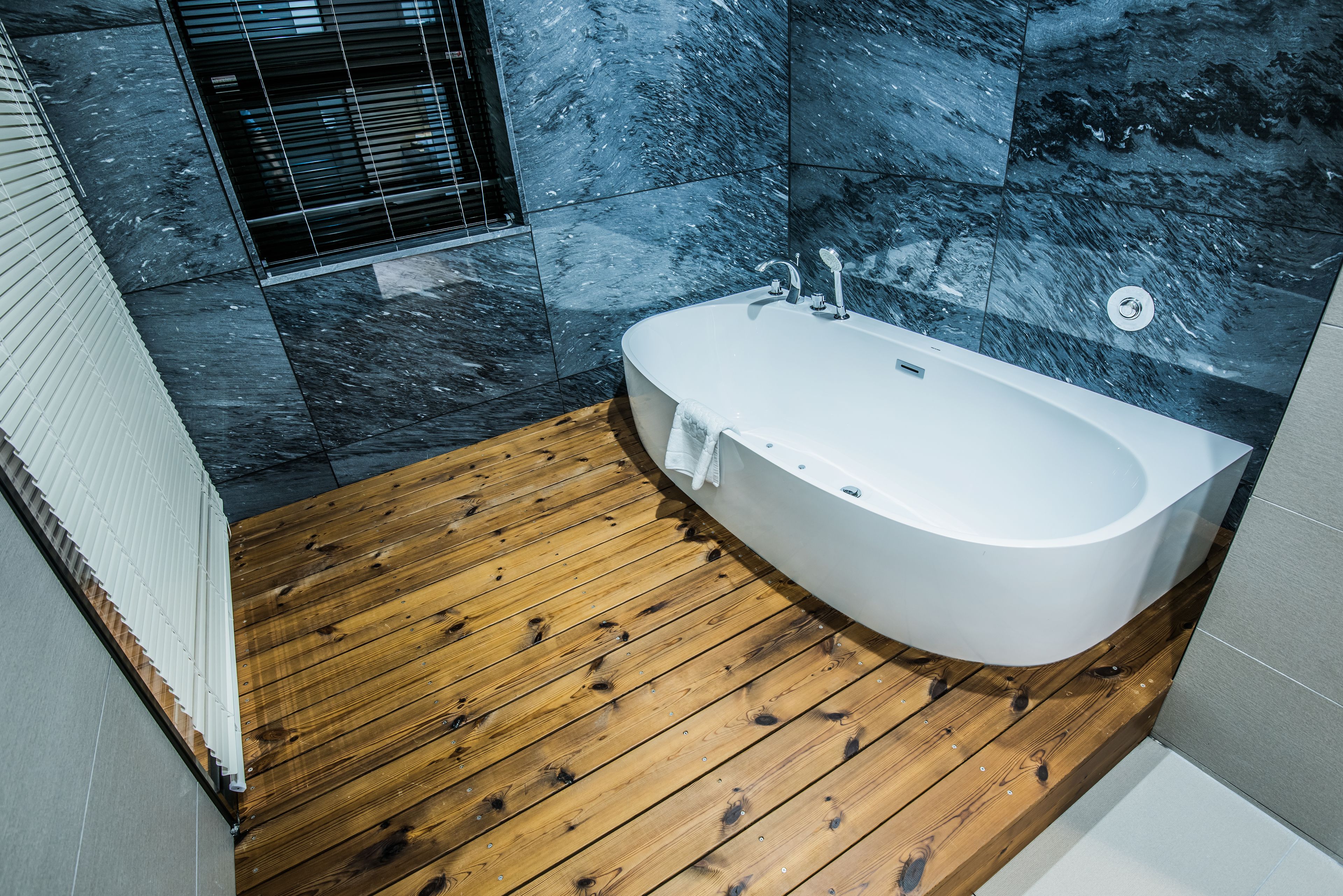 Un baño moderno con suelo de madera, una bañera independiente y paredes de mármol oscuro.