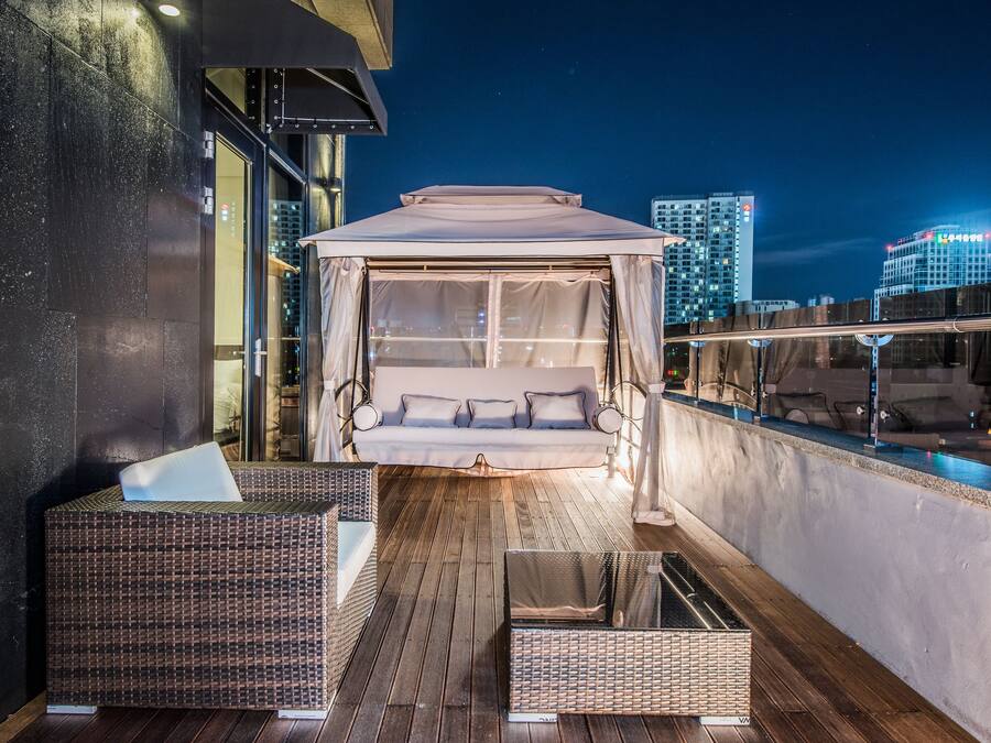 Un balcón con cama con dosel, mobiliario de mimbre y vista nocturna de la ciudad.
