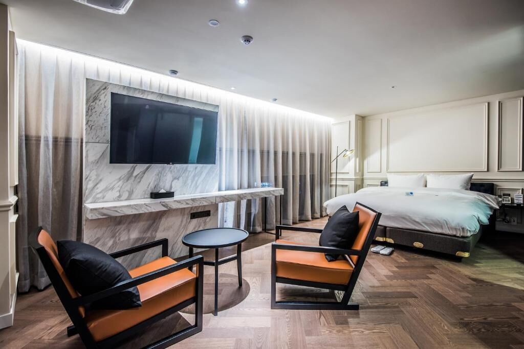 Une chambre d’hôtel moderne avec un grand lit, un téléviseur fixé sur un mur de marbre, une petite table ronde et deux chaises avec des coussins.