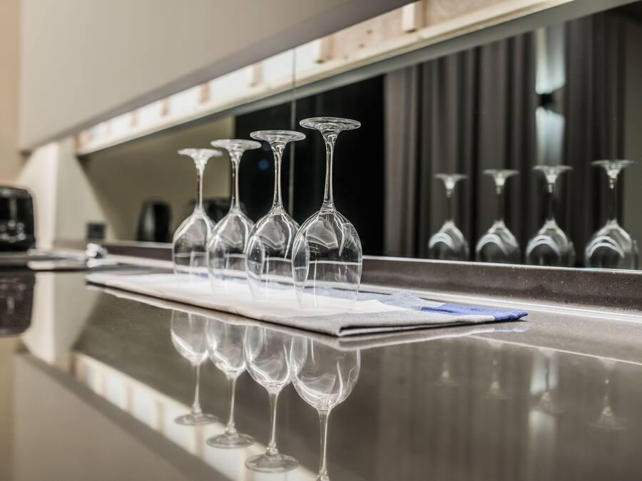 Una fila de copas de vino vacías sobre una encimera reflectante, con una máquina de café y un menú al fondo.