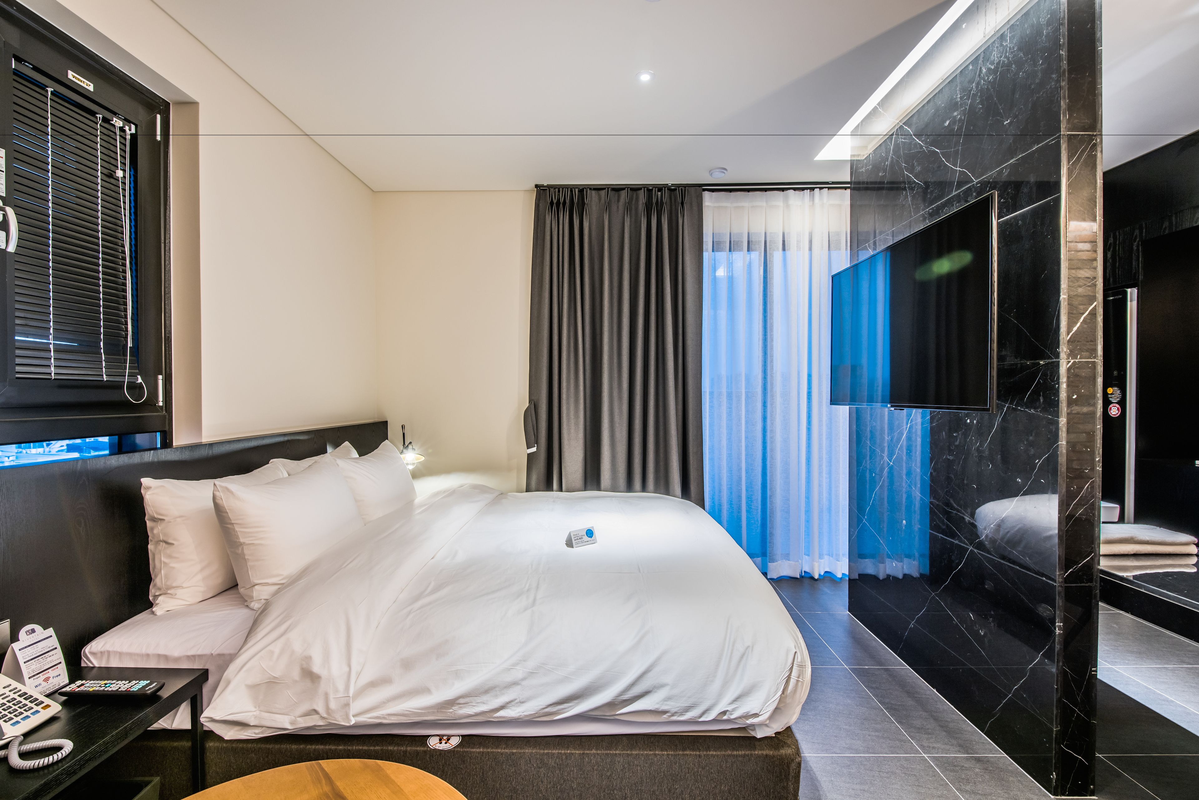 Ein modernes Hotelzimmer mit einem großen Bett, einem Nachttisch mit Telefon, einem Fernseher und Blick auf die Stadt durch das Fenster.