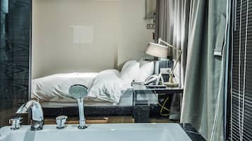 A modern hotel room with a large bathtub, a bed with white linens, a desk with a lamp, and a window with curtains.