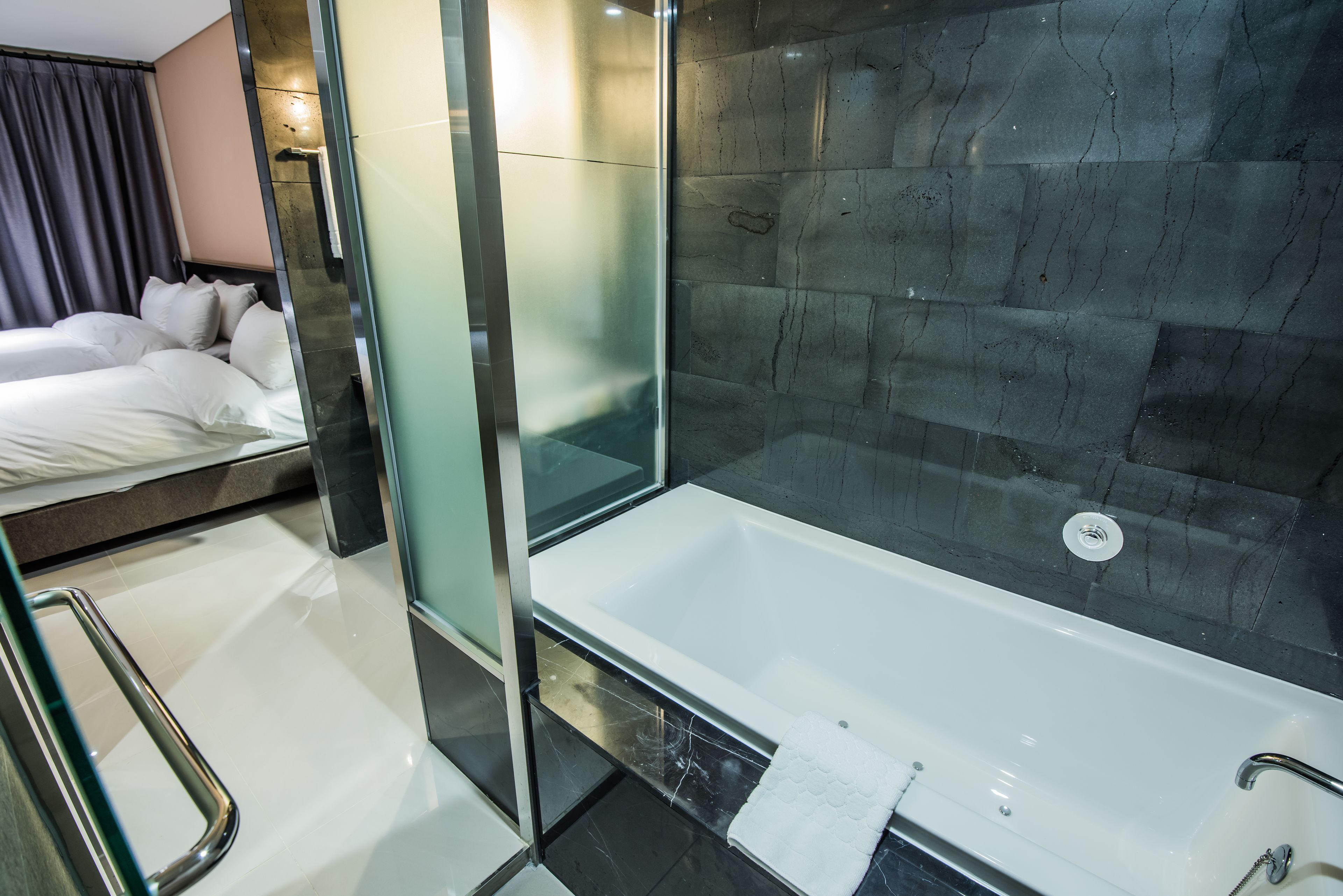 A modern bathroom with a glass-enclosed shower, a white bathtub, and a bed with white linens.