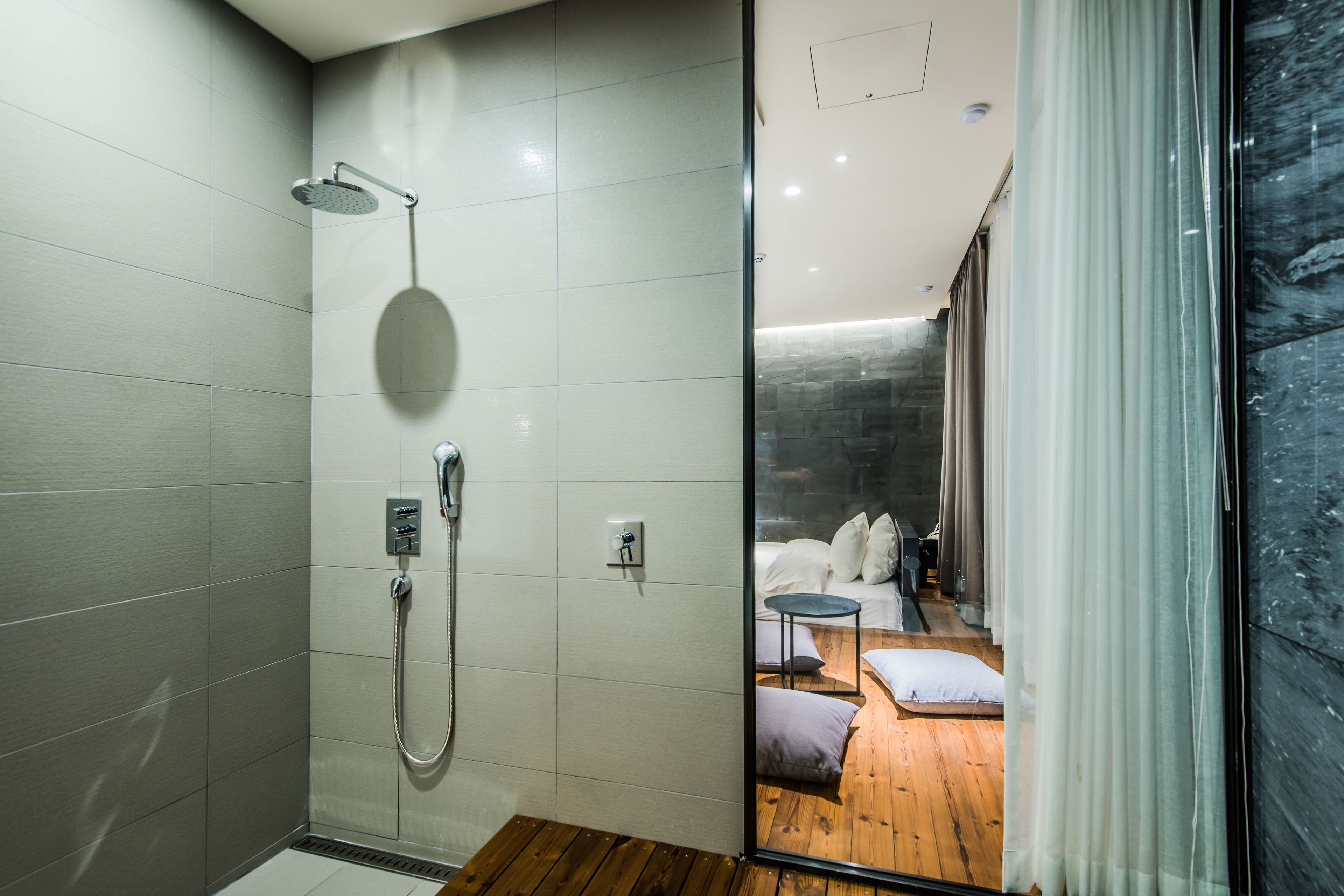 Un baño moderno con cabina de ducha de vidrio, suelo de madera y una habitación visible con una cama y una mesita.