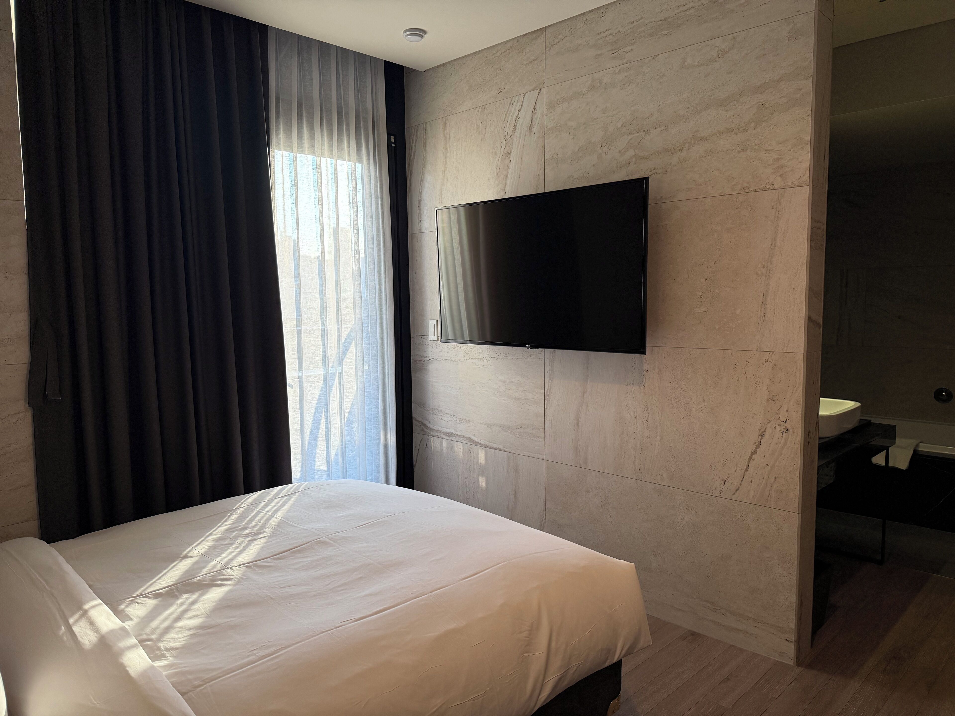 A modern hotel room with a large bed, a flat-screen TV mounted on the wall, and a bathroom visible through an open door.