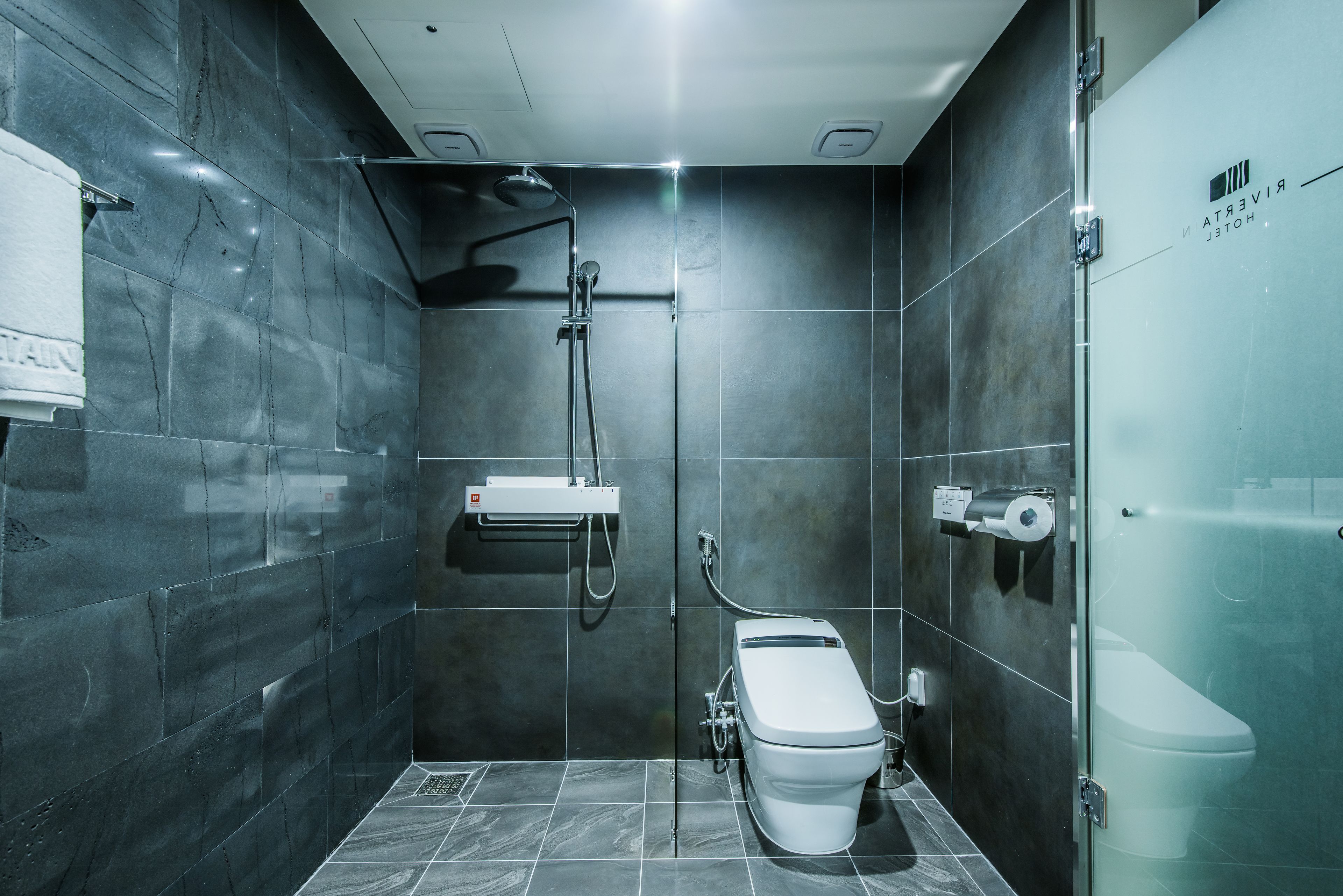 Un baño moderno con inodoro separado por mampara de vidrio, ducha con cabezal de ducha de mano y dispensador de toallas mural.