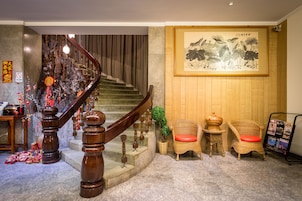 Una escalera con barandilla de madera, una obra de arte enmarcada en la pared, sillas de mimbre con cojines rojos y una mesita con una planta.