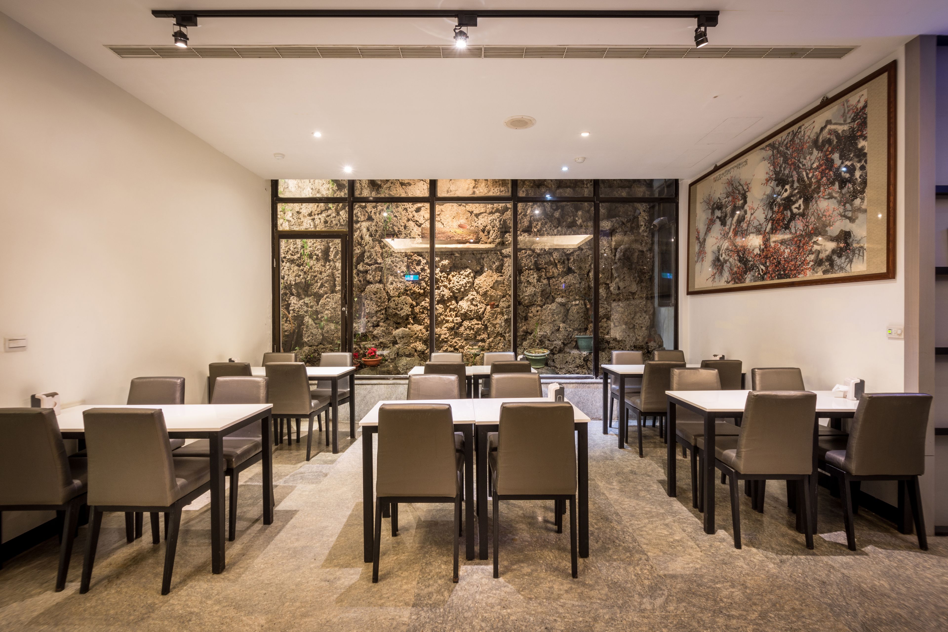 A conference room with tables and chairs, a large painting on the wall, and a view of a rocky wall through large windows.