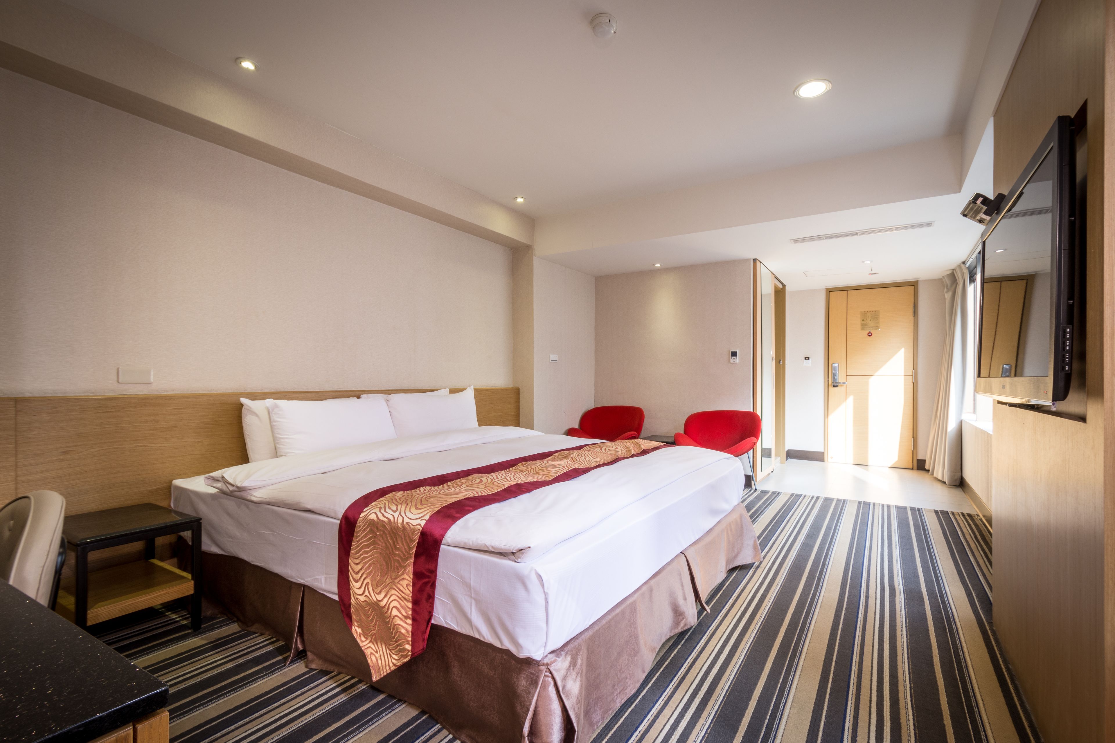 A hotel room with a large bed, a desk, two red chairs, and a TV on the wall.