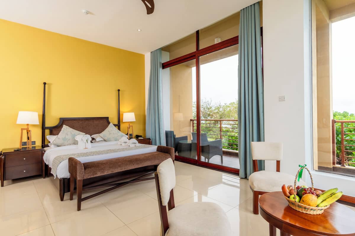 A hotel room with a bed, bedside tables, a dining table with fruit, a chair, and a balcony with a view.