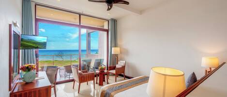 Camera d'hotel con un'ampia finestra vista oceano, un letto con vista, una scrivania con televisore e una poltrona.