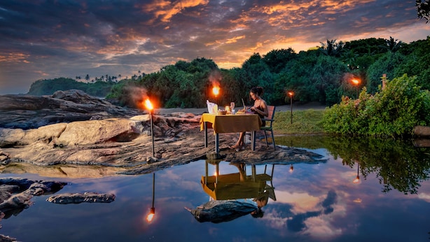A dining table with chairs and a person seated, set up on a rocky shore with a body of water in the foreground. - Sooriya Resort & Spa (Rekawa)