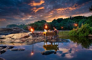 A dining table with chairs and a person seated, set up on a rocky shore with a body of water in the foreground. - Sooriya Resort & Spa (Rekawa)