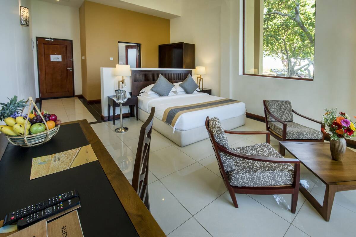 A hotel room with a bed, two chairs, a dining table, a fruit basket, a remote control, and a window with a view of trees.