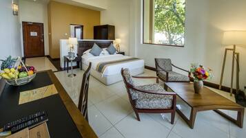 A hotel room with a bed, two chairs, a dining table, a fruit basket, a remote control, and a window with a view of trees.