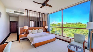 A spacious bedroom with a large bed, a sitting area with a chair, a table, and a view of lush greenery through large windows. - Sooriya Resort & Spa (Rekawa)