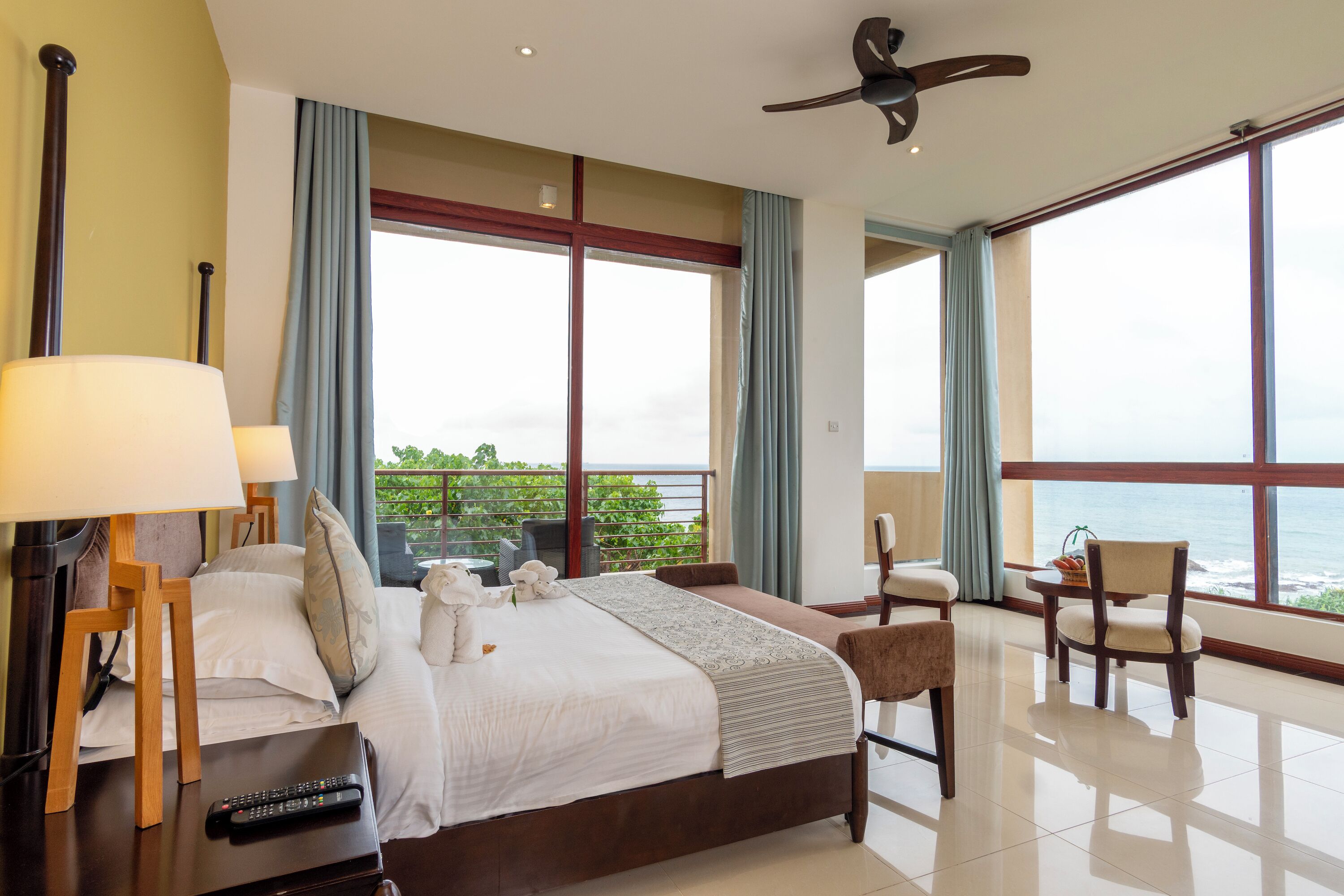 Quarto espaçoso com uma cama grande, um ventilador de teto e vista para o mar através de amplas janelas.