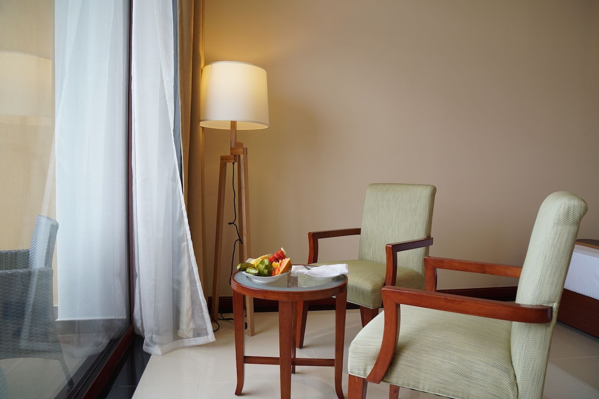A hotel room with a bed, a desk with a lamp, two chairs, and a small table with fruit.