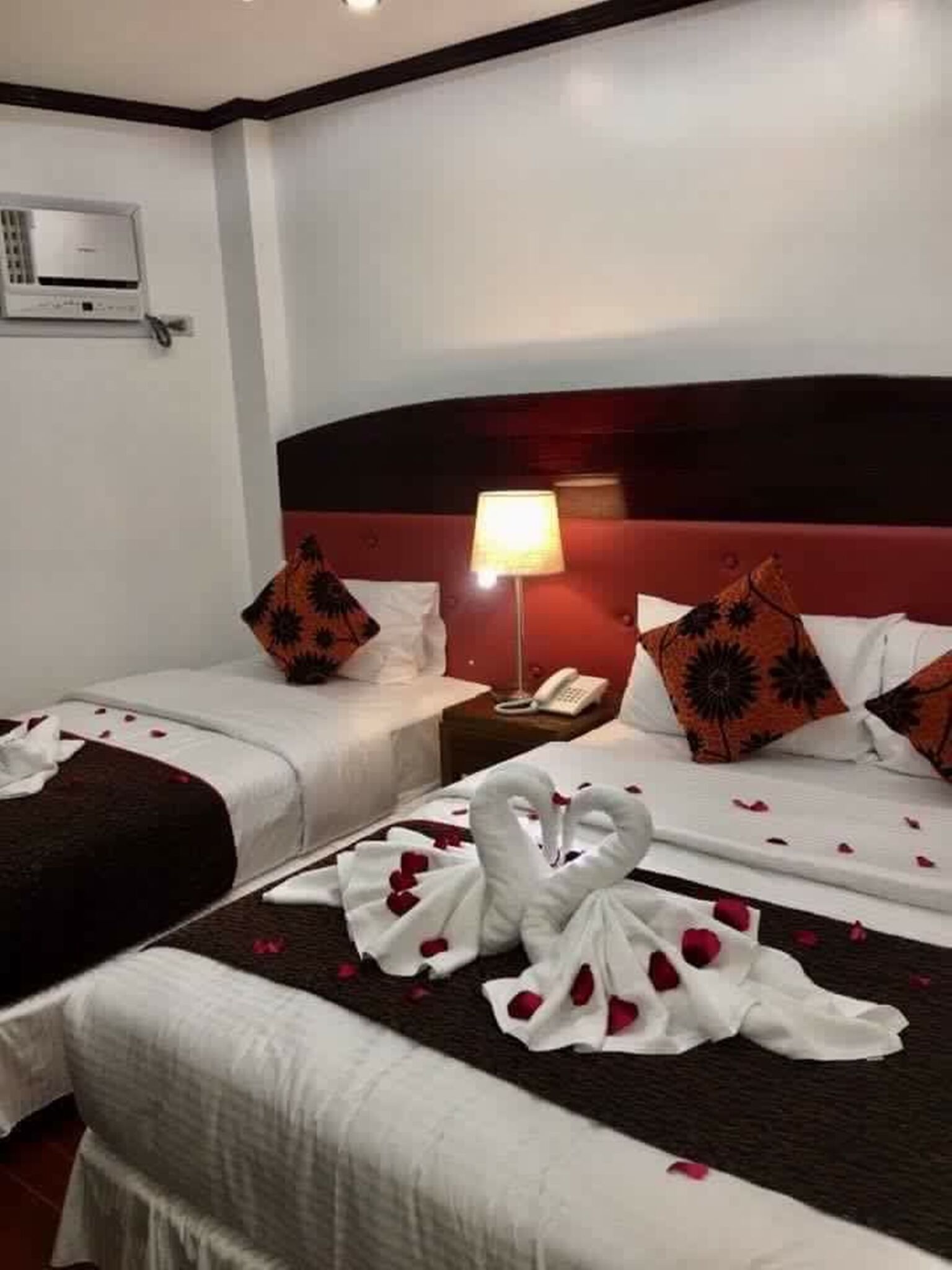 A hotel room with two beds, each with a red and black headboard, white bedding, and decorative pillows. There is a nightstand with a lamp and a phone.