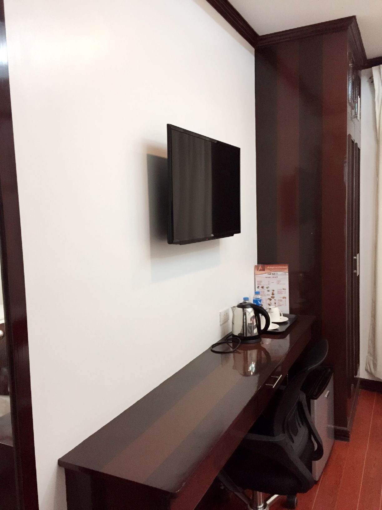 Habitación de hotel con televisor de pantalla plana montado en la pared, un escritorio de madera con una silla y una pequeña mesa con hervidor y tazas.
