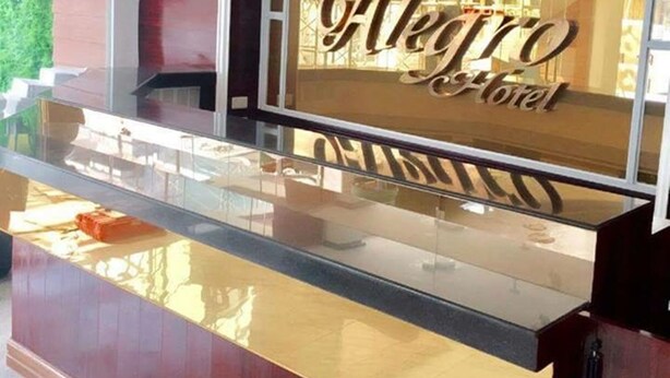 Uma receção com bancada de vidro e base de madeira, exibindo o nome 'Alegro Hotel' numa fonte decorativa.