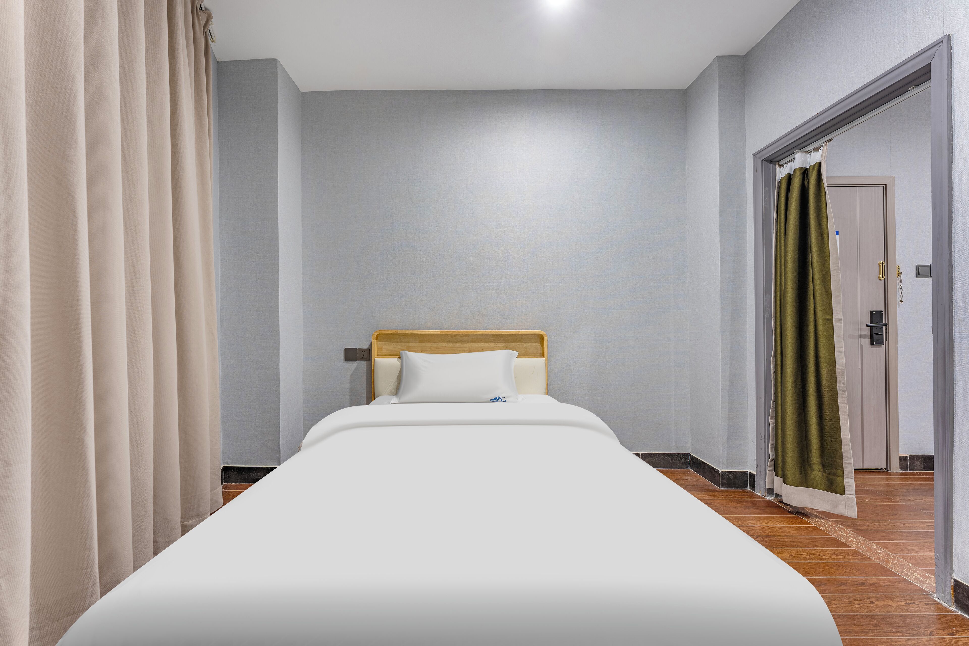 Una cama bien hecha con cabecera de madera, almohada blanca y cortina color beige a la izquierda.